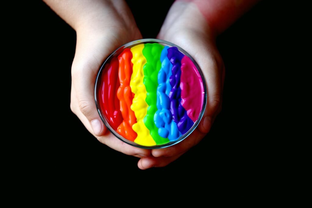 Close-up of hands holding a round container of rainbow-colored paint against a black background, symbolizing LGBTQ pride.